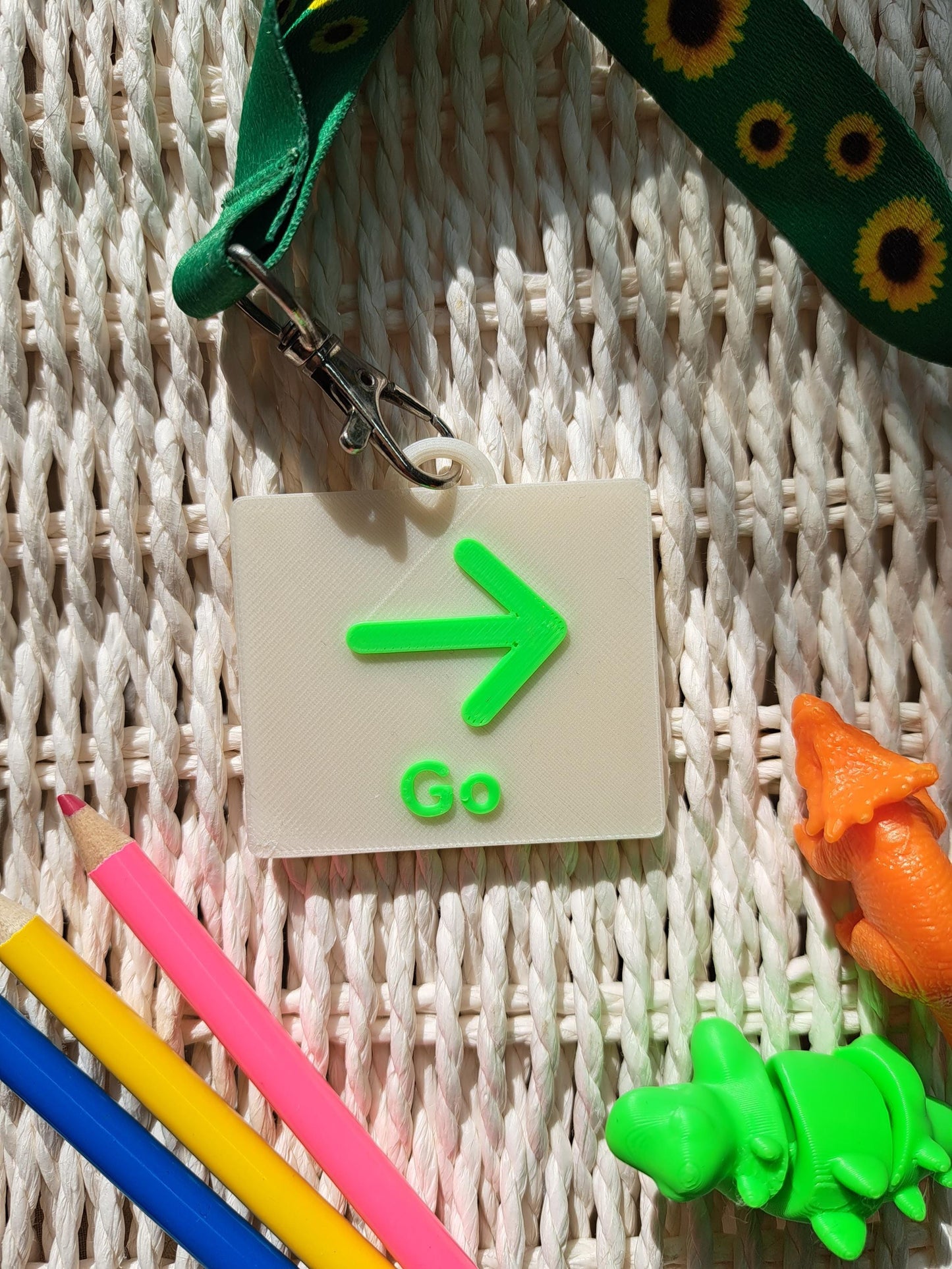 A 3D-printed "Stop" communication tag with a raised red hand symbol and bold red lettering, attached to a sunflower-patterned lanyard. The tag is placed on a textured white surface, surrounded by sensory-friendly tools, including colorful pencils and small dinosaur fidget toys. Designed as an autism communication aid for nonverbal children and individuals with speech delays.