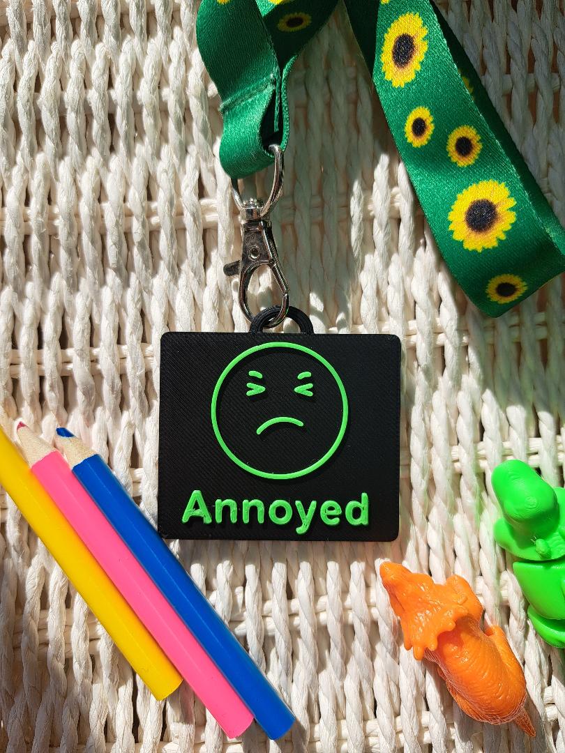 A black-colored 3D-printed communication tag with a raised green "Annoyed" face and the word "annoyed" below it. This visual aid tag is designed to support nonverbal communication, autism-friendly tools, and AAC needs.