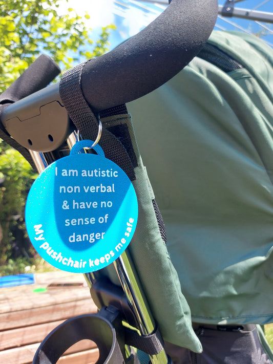A blue double-sided wheelchair tag attached to the handle of a green wheelchair, with text explaining the user's autism and non-verbal communication needs.