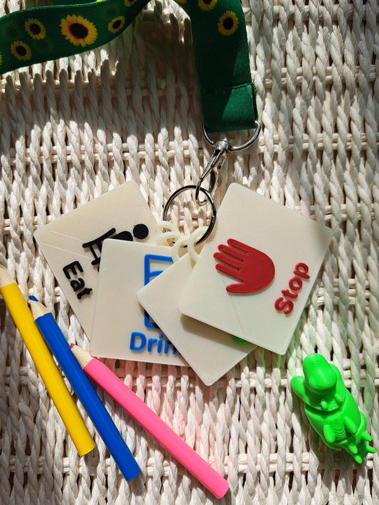A set of 3D-printed action communication tags, including “Stop” in red, “Go” in green, “Drink” in blue, and “Eat” in black, attached to a sunflower-patterned lanyard. Surrounding the tags are colorful pencils and a green dinosaur fidget toy.