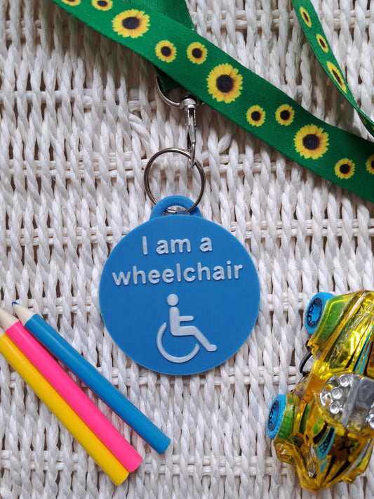 A blue, circular 3D-printed tag with white raised text reading "I am a wheelchair" and a wheelchair symbol, attached to a green sunflower lanyard with a metal clasp.
