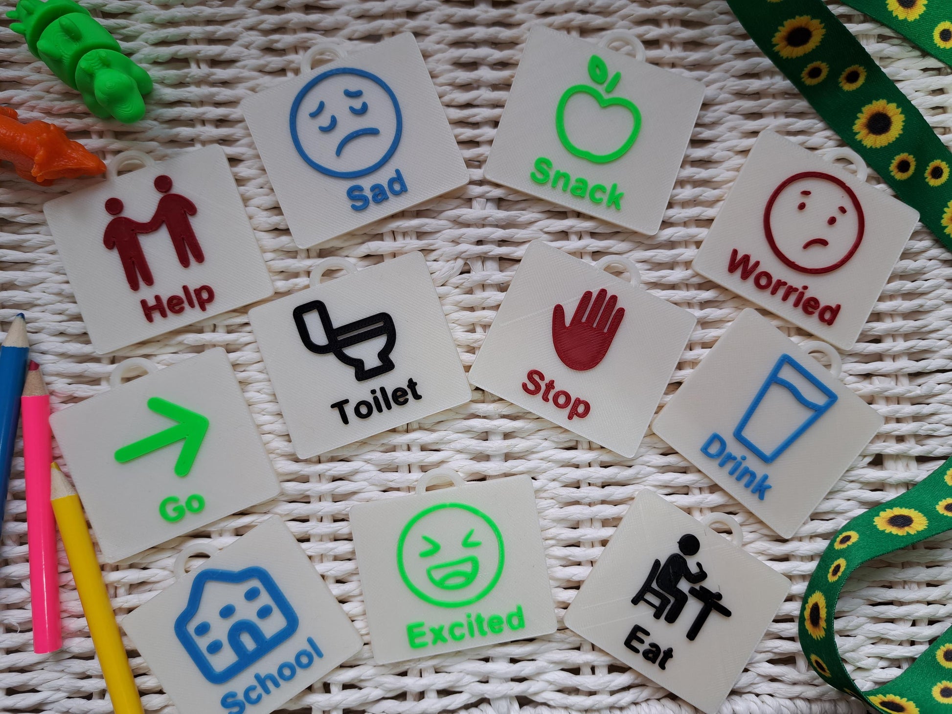 A set of 11 cream-colored 3D-printed communication tags with raised, colorful icons and text, arranged on a woven white surface. The tags display words Help, Sad, Snack, Worried, Go, Toilet, Stop, Drink, School, Excited, Eat in different colors.