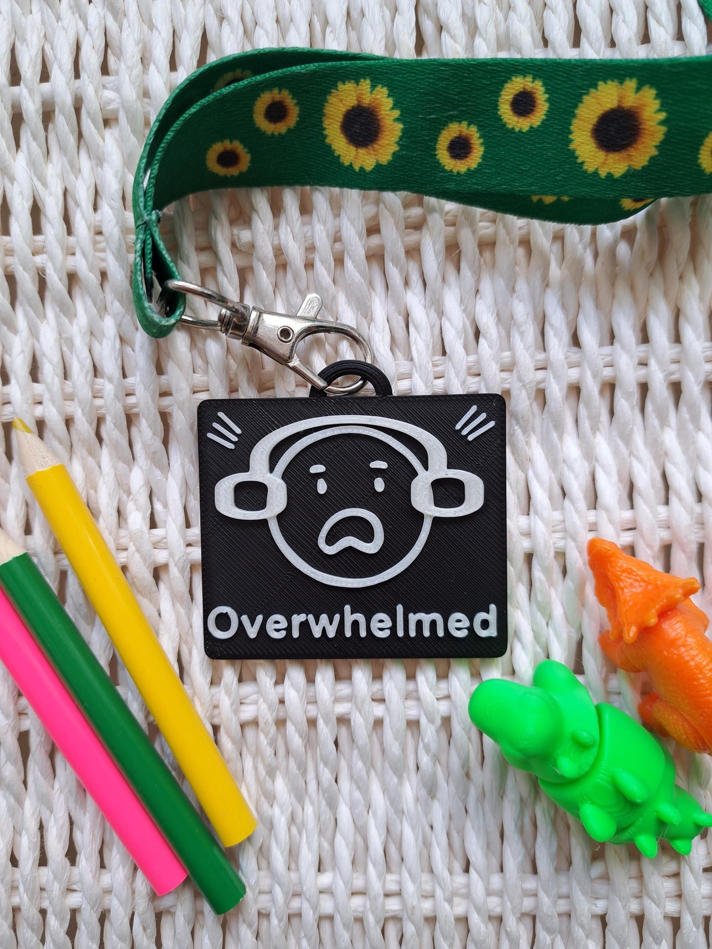 A black-colored 3D-printed communication tag with a raised white "overwhelmed" face with ear defenders on and the word "overwhelmed" below it. This visual aid tag is designed to support nonverbal communication, autism-friendly tools, and AAC needs.