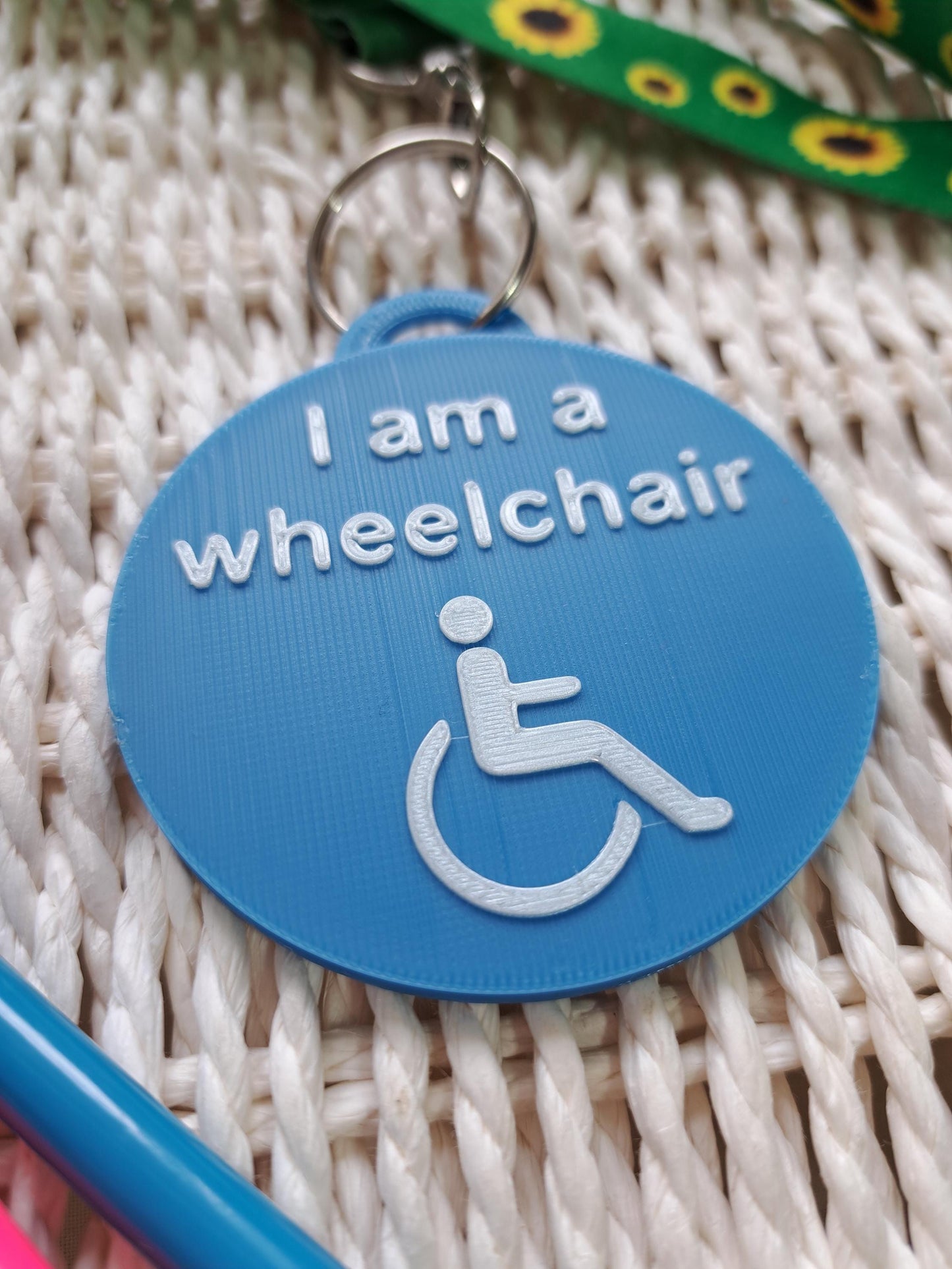 A blue, circular 3D-printed tag with white raised text reading "I am a wheelchair" and a wheelchair symbol, attached to a green sunflower lanyard with a metal clasp.