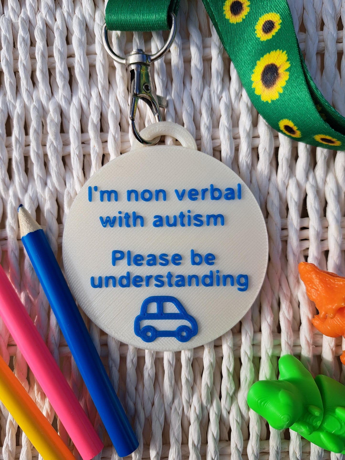 Autism ID Tag with 'I'm Non Verbal with Autism, Please Be Kind and Understanding' text, attached to a sunflower lanyard. Includes a blue car icon, surrounded by colorful pencils and sensory fidget toys. Ideal for autism awareness, backpacks, and lanyards.