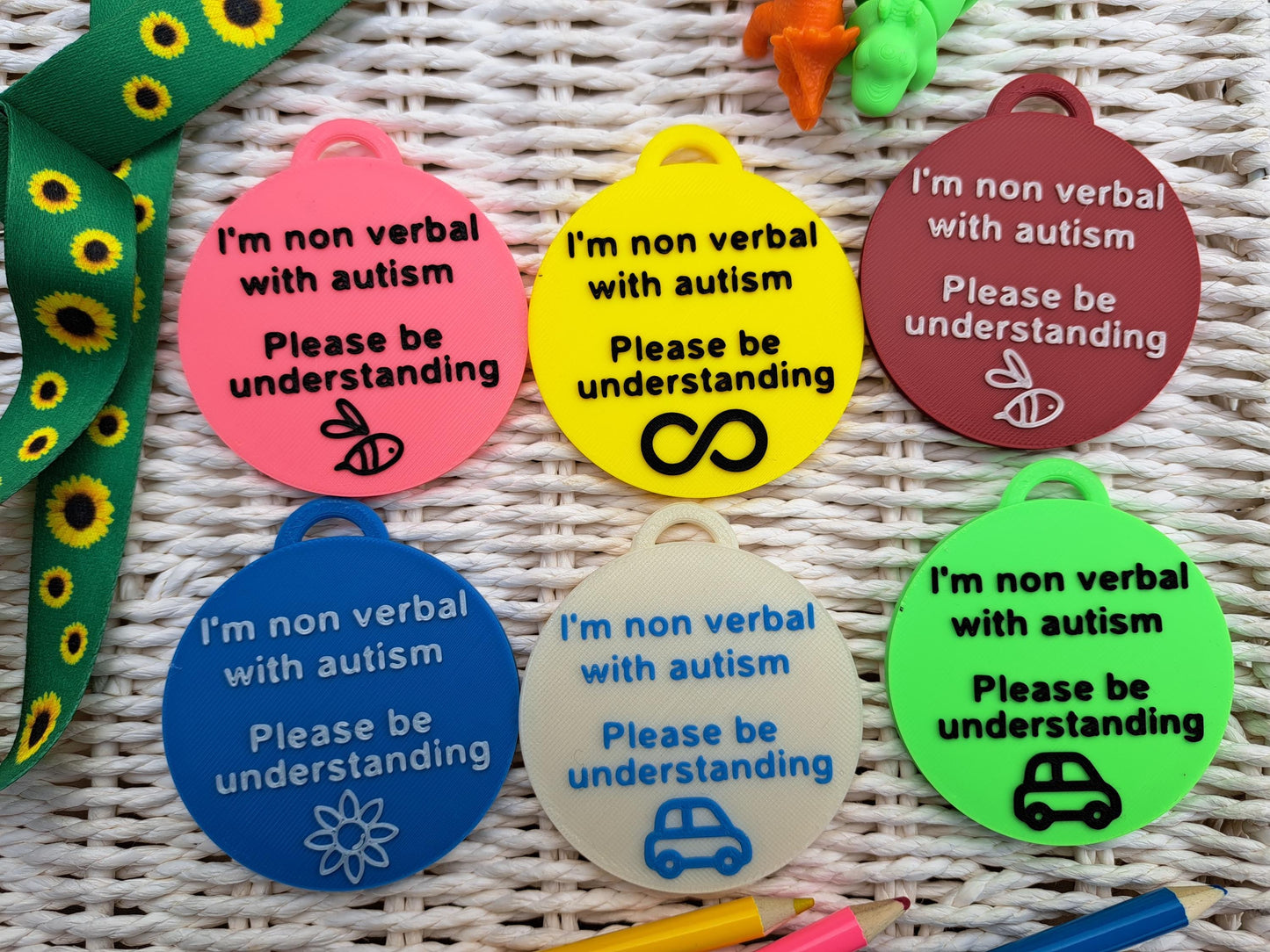 Set of six Autism ID Tags in yellow, green, pink, white, red, and blue, featuring 'I'm Non Verbal with Autism, Please Be Kind and Understanding' text. Each tag has a unique symbol: bee, infinity symbol, car, flower, and dinosaur. Ideal for autism awareness, backpacks, and lanyards.
