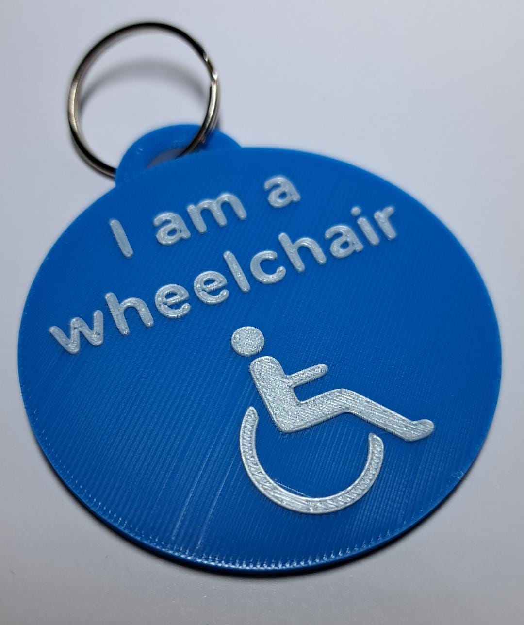 A blue, circular 3D-printed tag with white raised text reading "I am a wheelchair" and a wheelchair symbol, attached to a green sunflower lanyard with a metal clasp.