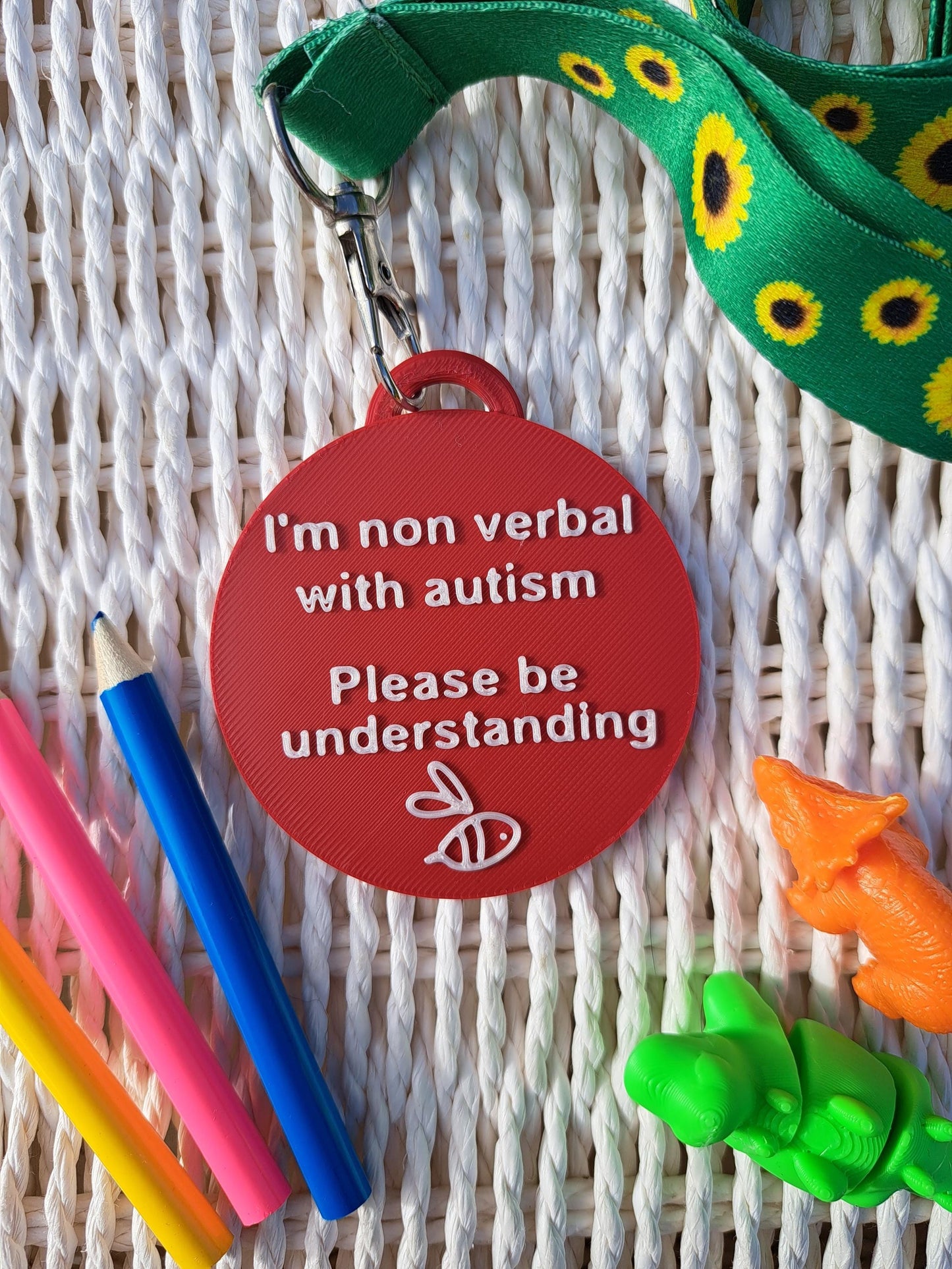 Autism ID Tag with 'I'm Non Verbal with Autism, Please Be Kind and Understanding' text, attached to a sunflower lanyard. Includes a white bee icon, surrounded by colorful pencils and sensory fidget toys. Ideal for autism awareness, backpacks, and lanyards.
