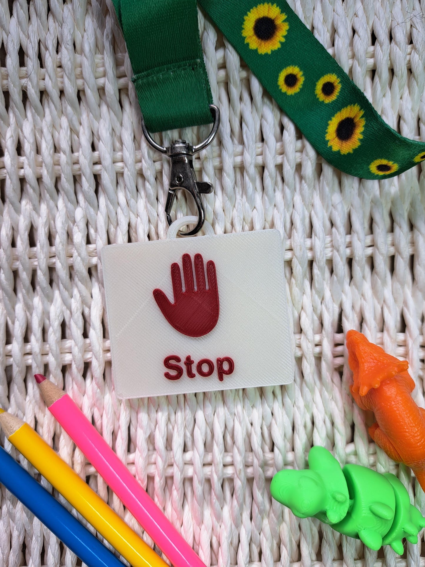 A 3D-printed "Drink" communication tag with a raised blue cup symbol and bold blue lettering, attached to a sunflower-patterned lanyard. The tag is placed on a textured white surface, surrounded by sensory-friendly tools, including colorful pencils and small dinosaur fidget toys. Designed as an autism communication aid for nonverbal children and individuals with speech delays.