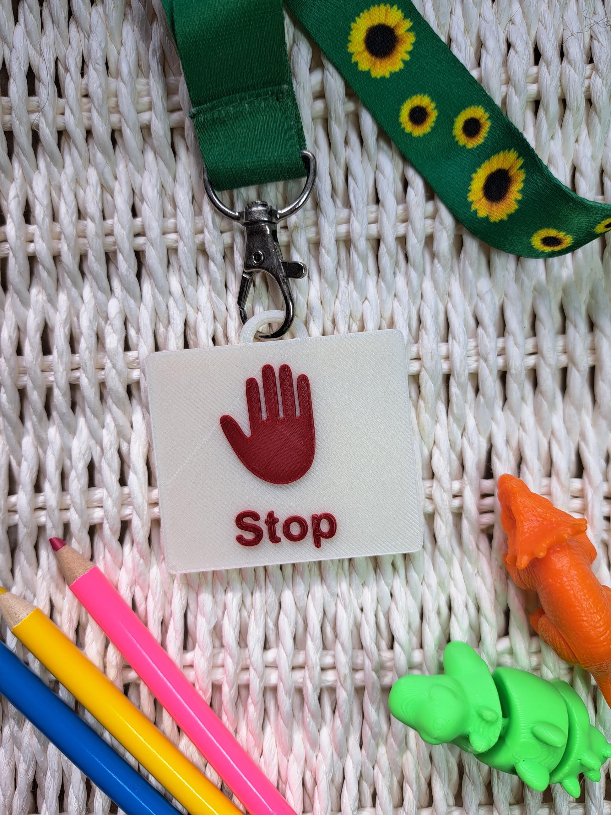 A 3D-printed "Drink" communication tag with a raised blue cup symbol and bold blue lettering, attached to a sunflower-patterned lanyard. The tag is placed on a textured white surface, surrounded by sensory-friendly tools, including colorful pencils and small dinosaur fidget toys. Designed as an autism communication aid for nonverbal children and individuals with speech delays.