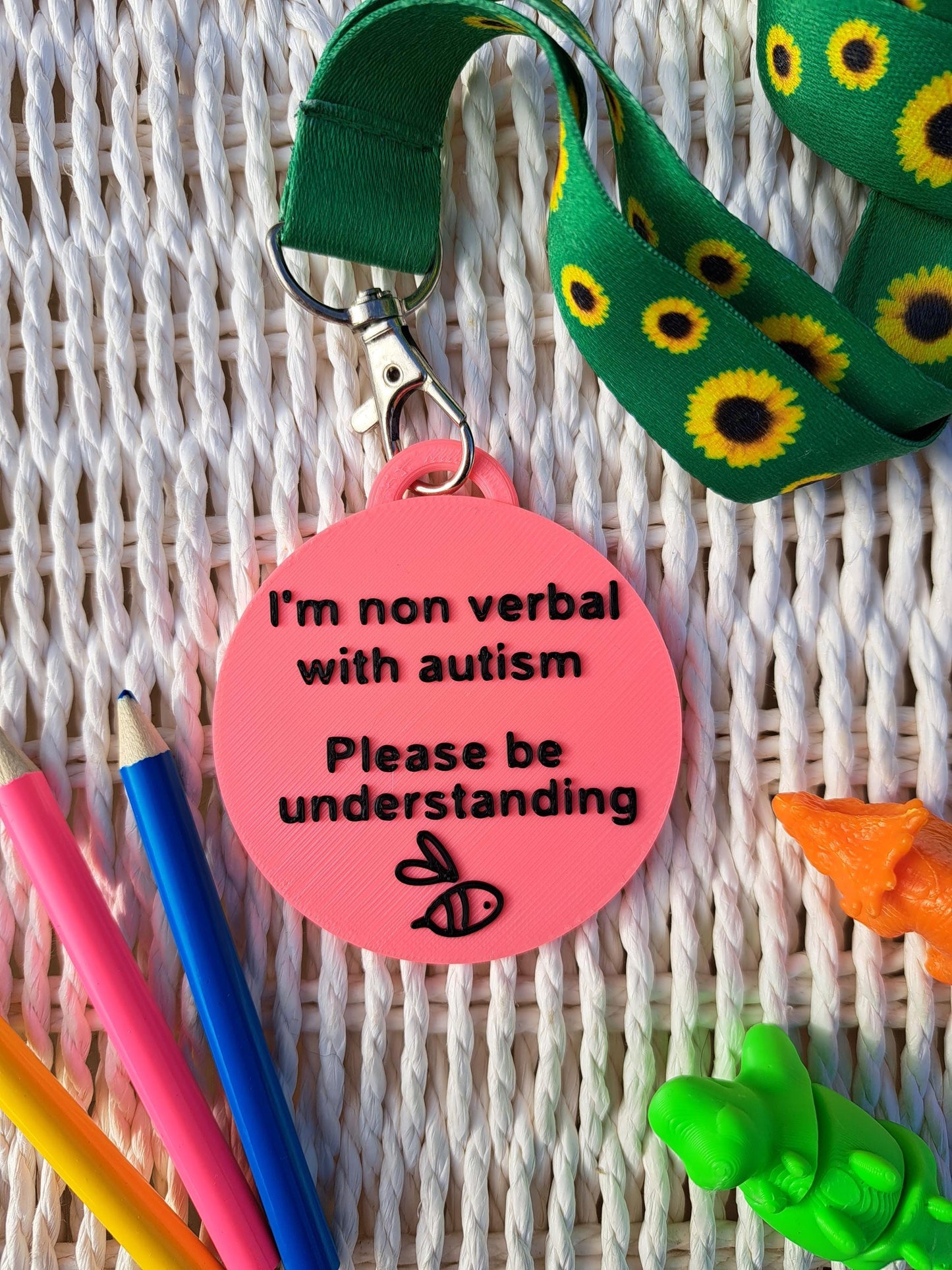 Autism ID Tag with 'I'm Non Verbal with Autism, Please Be Kind and Understanding' text, attached to a sunflower lanyard. Includes a black bee icon, surrounded by colorful pencils and sensory fidget toys. Ideal for autism awareness, backpacks, and lanyards.
