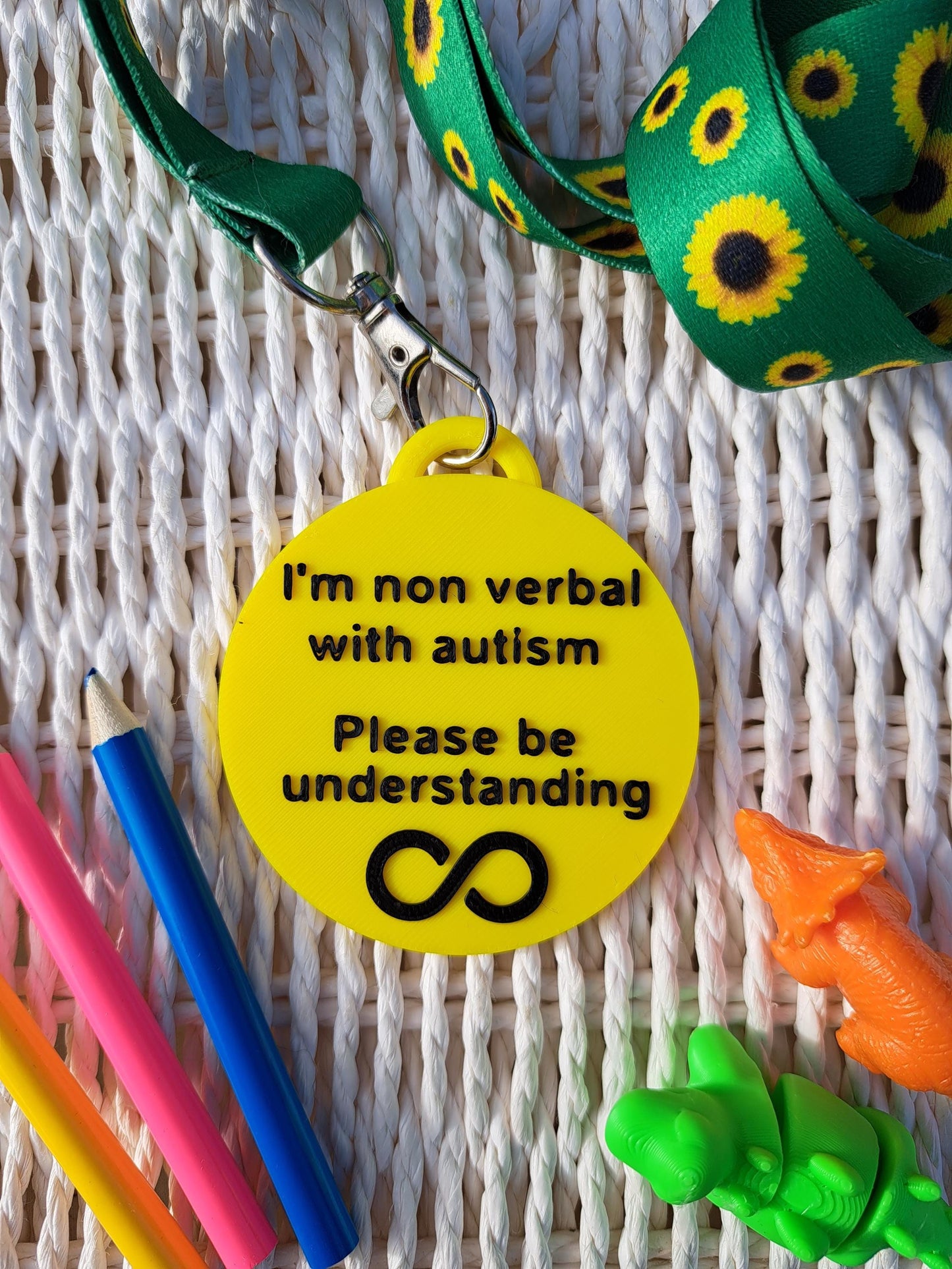 Autism ID Tag with 'I'm Non Verbal with Autism, Please Be Kind and Understanding' text, attached to a sunflower lanyard. Includes a black infinity icon, surrounded by colorful pencils and sensory fidget toys. Ideal for autism awareness, backpacks, and lanyards.