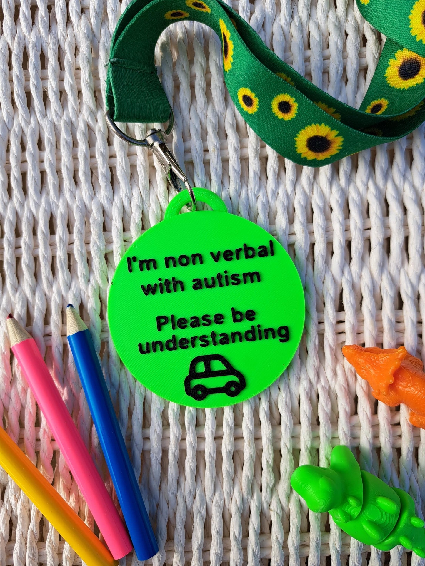 Autism ID Tag with 'I'm Non Verbal with Autism, Please Be Kind and Understanding' text, attached to a sunflower lanyard. Includes a black car icon, surrounded by colorful pencils and sensory fidget toys. Ideal for autism awareness, backpacks, and lanyards.