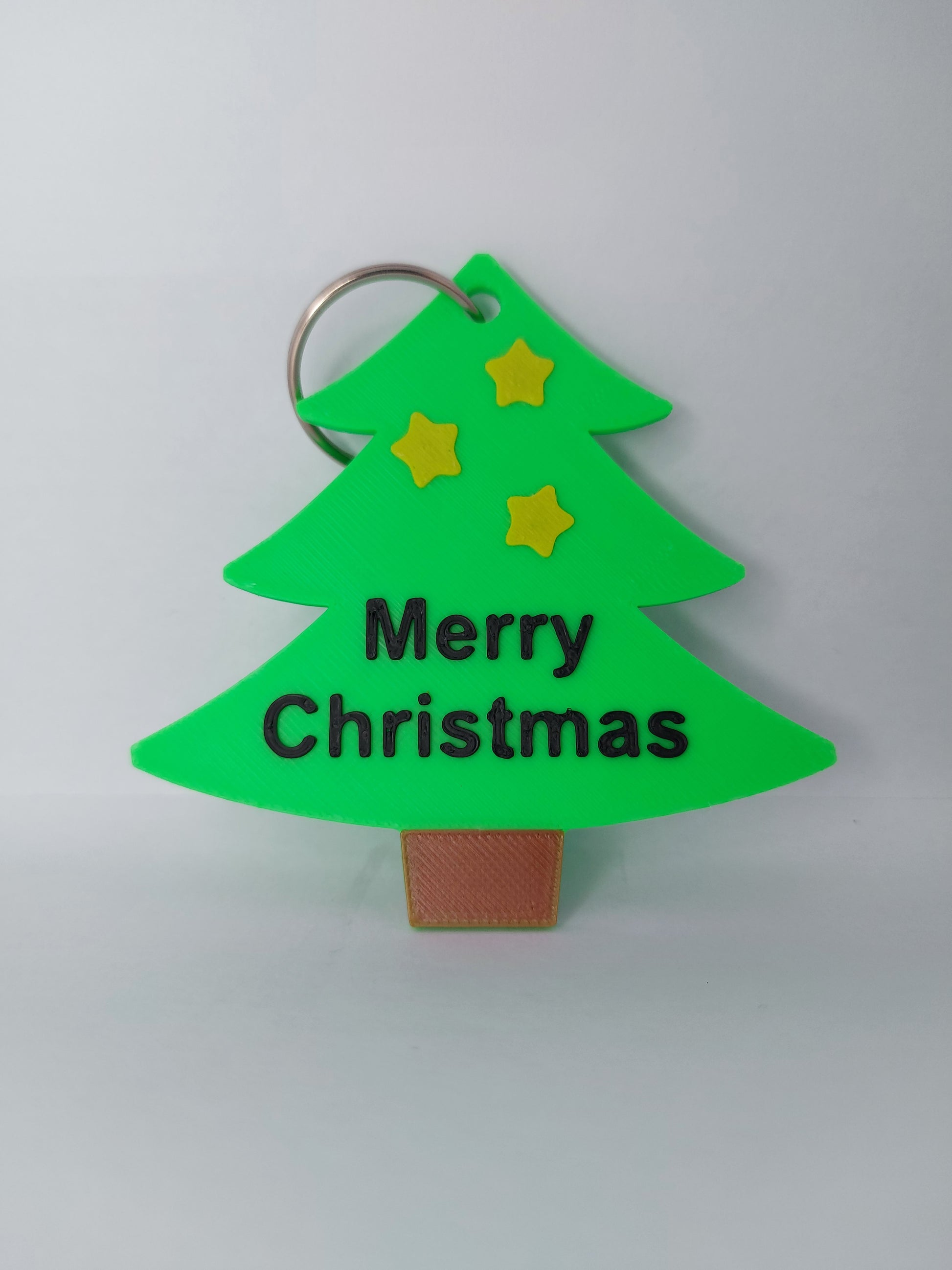 Green Christmas tree-shaped keychain with 'Merry Christmas' text and stars on a light gray background