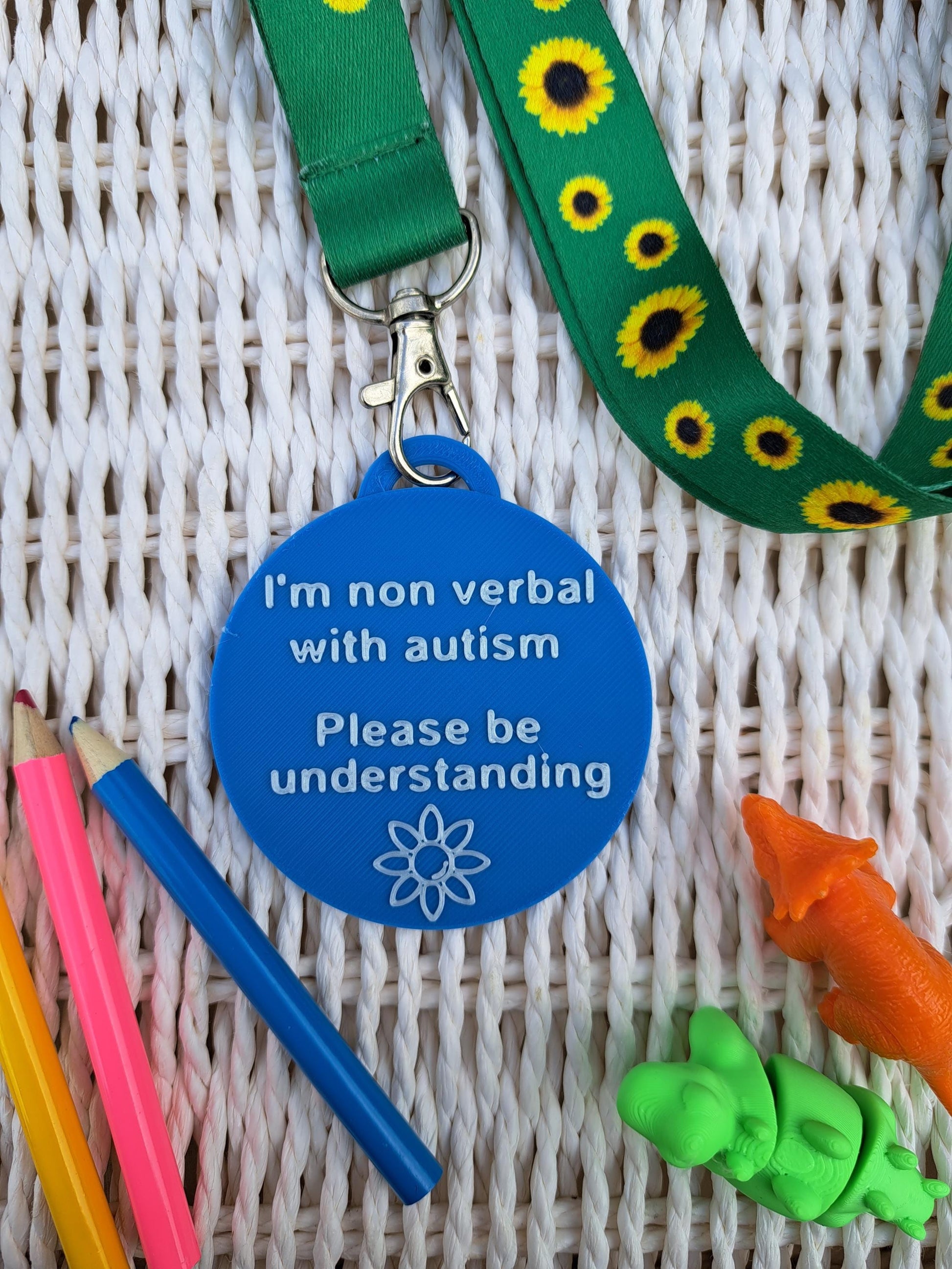 Autism ID Tag with 'I'm Non Verbal with Autism, Please Be Kind and Understanding' text, attached to a sunflower lanyard. Includes a white flower icon, surrounded by colorful pencils and sensory fidget toys. Ideal for autism awareness, backpacks, and lanyards.